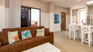 A modern living room with a brown sofa, two decorative pillows, a wooden coffee table, and a kitchen area with a white bar and stools.