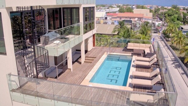 A rooftop pool with lounge chairs and a glass balustrade, overlooking a cityscape.