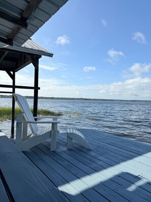 Une terrasse en bois avec deux chaises Adirondack blanches, un espace abrité et une vue sur l’eau et le rivage au loin.