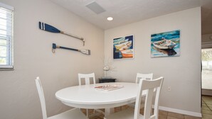 A dining area with a white table and chairs, two paintings of boats on the wall, and a paddle on the wall.
