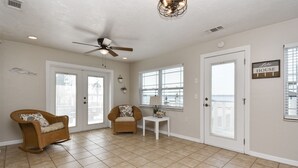 A spacious living room with a ceiling fan, two doors, a window with blinds, and a small table with a lamp.