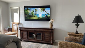 Ein Wohnzimmer mit an der Wand montiertem Fernseher, einem Holzschrank, Korbmöbeln und einer Lampe.