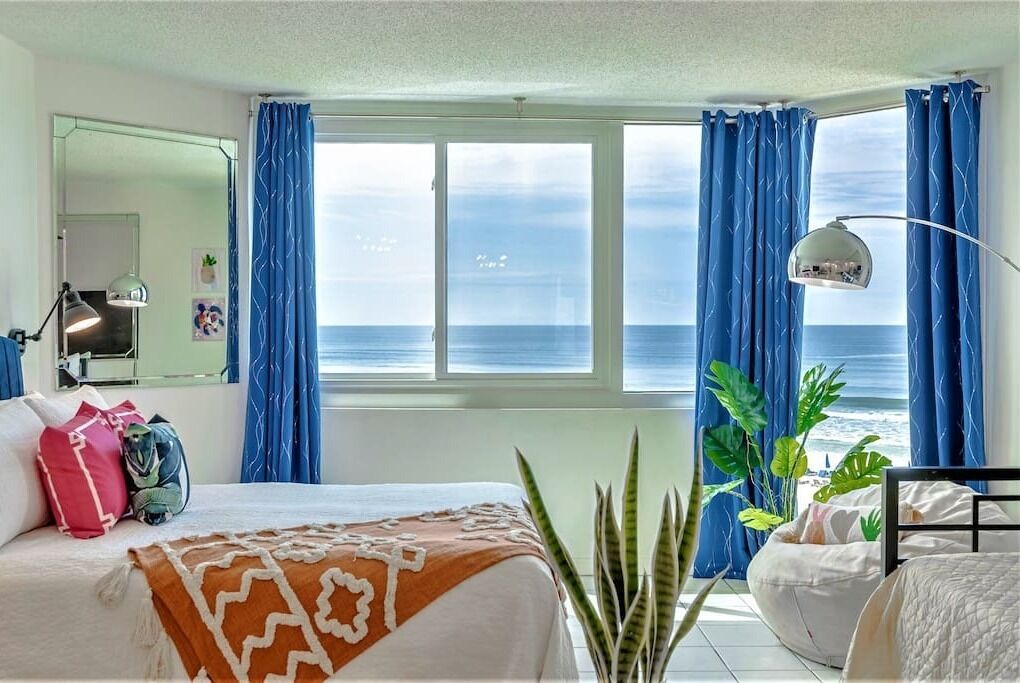 A bedroom with a bed, a desk, a chair, a mirror, and a view of the ocean.