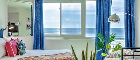 A bedroom with a bed, a desk, a chair, a mirror, and a view of the ocean.