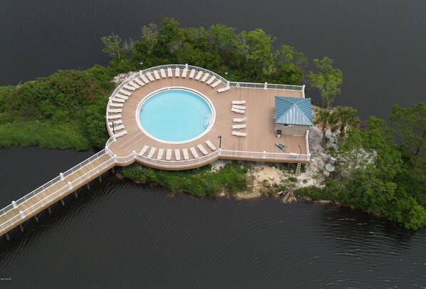 Vista aérea de una piscina circular rodeada por una terraza de madera con sillas de descanso, un pequeño edificio con techo azul y un puente de madera que conecta con tierra firme.