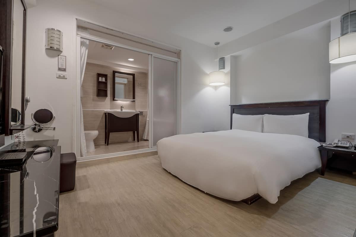 Habitación de hotel con cama, mesita de noche, lámpara y baño con lavabo y espejo.
