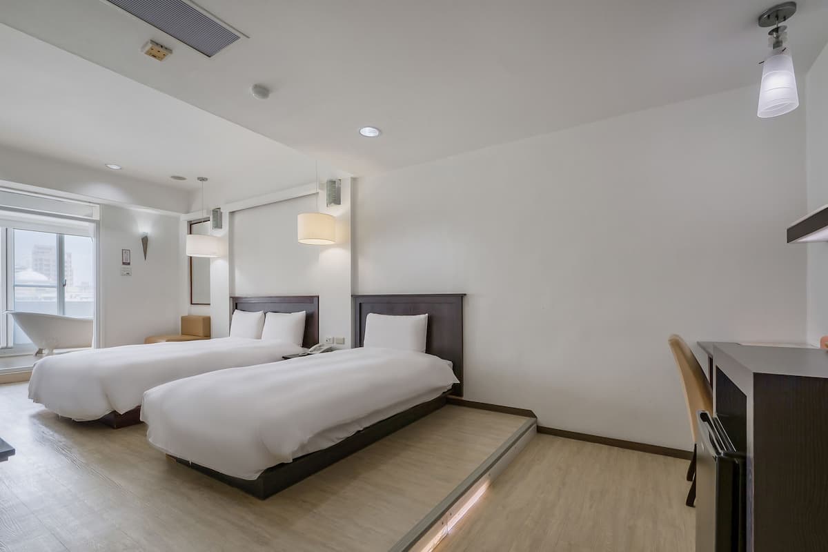 Habitación de hotel con dos camas, un escritorio, una silla, una ventana y baño.