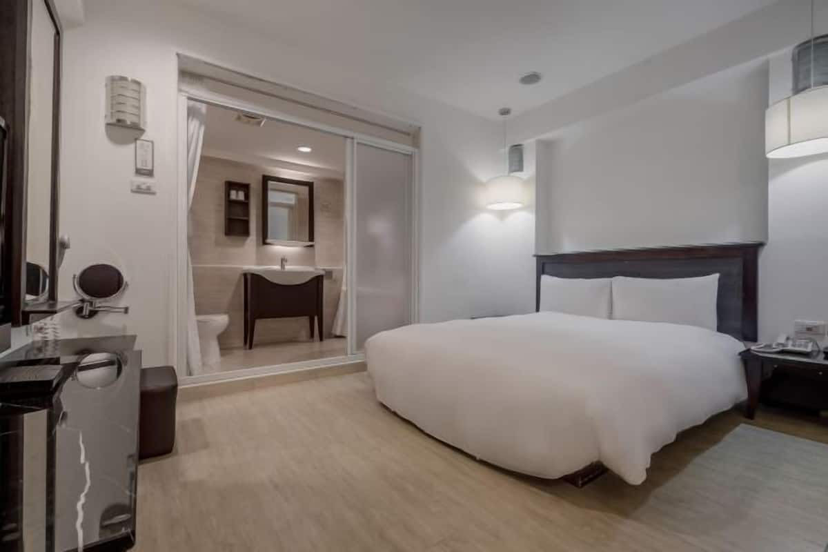 Habitación de hotel con cama, escritorio, silla, espejo y baño visible a través de una puerta abierta.