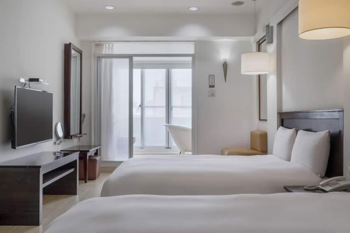 Habitación de hotel con dos camas, televisión, escritorio y baño visible a través de la puerta de cristal.