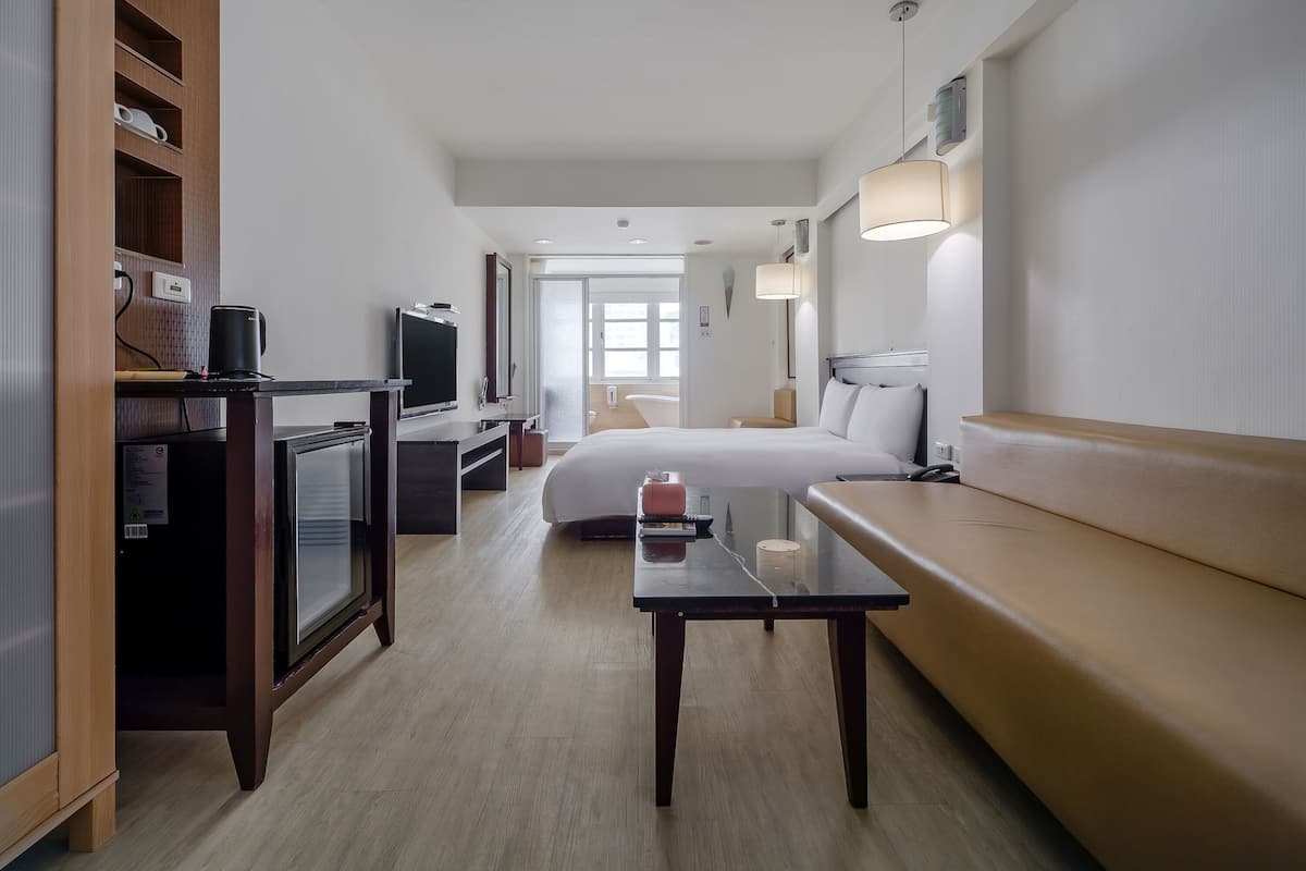 Habitación de hotel con cama, televisor, escritorio, sofá y baño visible a través de una puerta abierta.