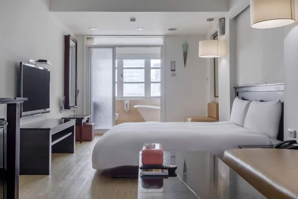 Una habitación de hotel moderna con una cama, un escritorio, un televisor y un baño visible a través de la puerta.