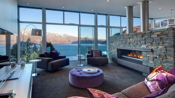 A modern living room with a stone fireplace, large windows, and a view of mountains and a lake. - Villa Matau | Gas Fire, Lake Views, A/C, BBQ (Queenstown)