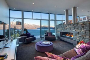A modern living room with a stone fireplace, large windows, and a view of mountains and a lake. - Villa Matau | Gas Fire, Lake Views, A/C, BBQ (Queenstown)