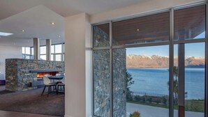 A modern interior with a stone fireplace, a dining area, and large windows overlooking a lake and mountains. - Villa Matau | Gas Fire, Lake Views, A/C, BBQ (Queenstown)