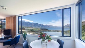 A modern room with a large window, a round table with chairs, a TV, and a plant. - Highlands Lodge – 5BR Luxury, Lake Views & BBQ (Queenstown)