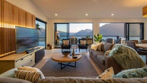 A modern living room with a large flat-screen TV, comfortable seating, a dining area, and a view of mountains through large windows. - Highlands Lodge – 5BR Luxury, Lake Views & BBQ (Queenstown)