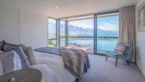 A bedroom with a large bed, a chair, and a view of a lake and mountains through large windows. - Highlands Lodge – 5BR Luxury, Lake Views & BBQ (Queenstown)