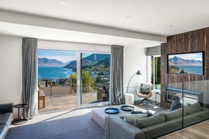 A modern living room with a large window, a sofa, a TV, and a balcony with a view of mountains and a lake. - Villa Habeke | Lake Views, Gas Fire, A/C, BBQ (Queenstown)