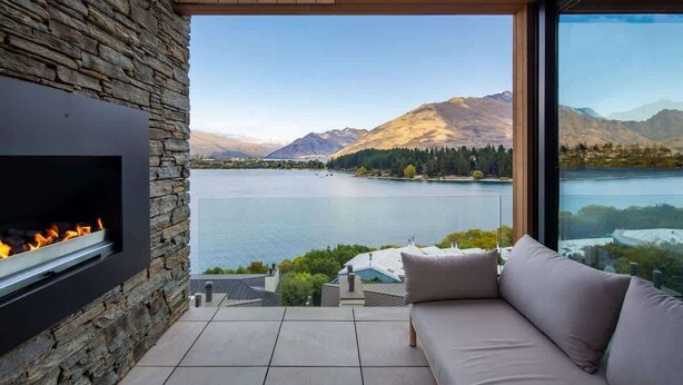 A modern outdoor seating area with a stone wall, a built-in fireplace, and a view of a lake and mountains. - Lakehouse 2 – Parking, Fireplace, AC, Lake Views (Queenstown)