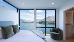 A bedroom with a large bed, a chair, and a view of a lake and mountains through large windows. - Lakehouse 2 – Parking, Fireplace, AC, Lake Views (Queenstown)