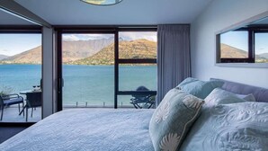 A bedroom with a large bed, a view of a lake and mountains, and a balcony with a table and chairs. - Kikorangi | Lake View, BBQ, A/C & Free Parking (Queenstown)