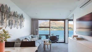 A modern living room with a sofa, dining table, and chairs, a flat-screen TV, and a large window with a view of a lake and hills. - Kikorangi | Lake View, BBQ, A/C & Free Parking (Queenstown)