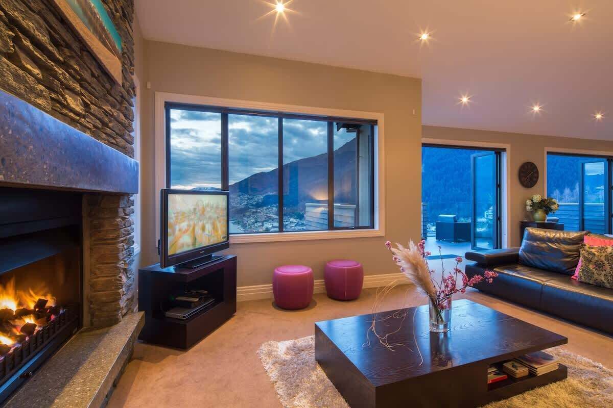A modern living room with a fireplace, a large window with a mountain view, a flat-screen TV, and a sofa.