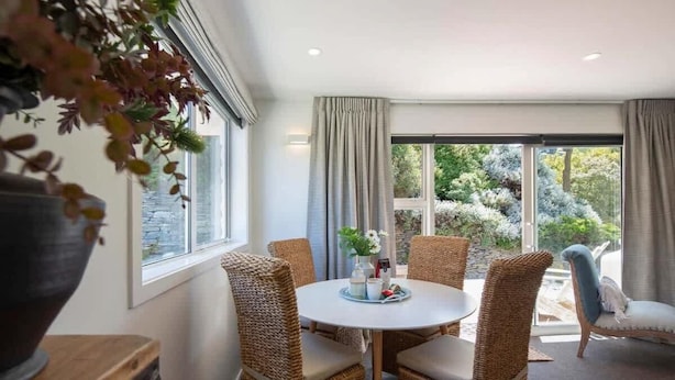 A dining area with wicker chairs, a round table, and a view of the outdoors through large windows. - Hillside Villa 4 – Modern 2BR Walk to Town & A/C (Queenstown)