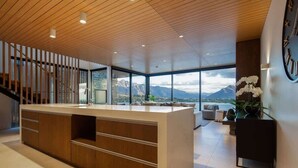 A modern interior with a wooden ceiling, a central island with drawers, a staircase, and a view of mountains through large windows. - Lakehouse 4 – Parking, Fireplace, Lake Views (Queenstown)