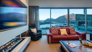 A modern living room with a red sofa, a black armchair, a fireplace, and a large window offering a view of mountains and a body of water. - Aurora Lodge– Hot Tub, Fireplace & Lake Views, BBQ (Queenstown)