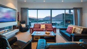 A modern living room with a large TV, comfortable seating, a fireplace, and a scenic view. - Aurora Lodge– Hot Tub, Fireplace & Lake Views, BBQ (Queenstown)