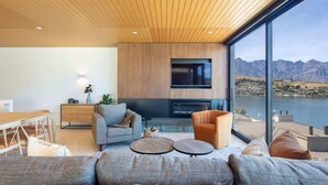 Ein modernes Wohnzimmer mit großem Fernseher, bequemen Sitzmöglichkeiten und Blick auf einen See und Berge.