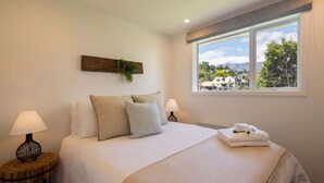 A neatly made bed with pillows and a folded towel, two bedside lamps, a wall-mounted plant, and a window with a view of a house and trees. - Hillside Villa 3 – Modern 2BR Walk to Town & A/C (Queenstown)