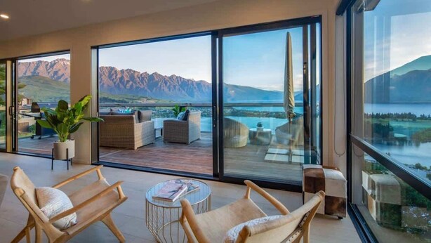 A modern interior with large windows, a balcony with outdoor furniture, and a view of mountains and water. - Highlands House – Lake Views, BBQ & Luxury Stay (Queenstown)