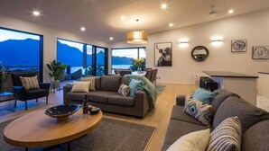 A modern living room with a sofa, coffee table, and dining area, large windows with a mountain view, and a pendant light. - Highlands House – Lake Views, BBQ & Luxury Stay (Queenstown)