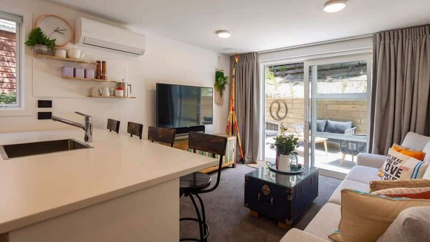 A modern kitchen with a central island, a dining area with chairs, a flat-screen TV, and a seating area with a sofa and cushions. - Hillside Villa 2 – Modern 2BR Walk to Town & A/C (Queenstown)