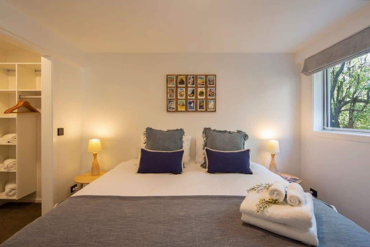 A hotel room with a bed, two bedside lamps, a wall-mounted photo collage, and a window with a view of trees.