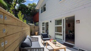 A patio with wicker furniture, a wooden table, and a white building with a sliding glass door. - Hillside Villa 2 – Modern 2BR Walk to Town & A/C (Queenstown)