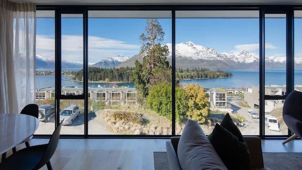 A room with a large window offering a view of a lake, mountains, and a parking area. - Villa Pukaki | Gas Fire, Free Parking, Lake Views (Queenstown)