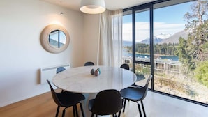 A modern dining area with a round table, black chairs, a hanging light fixture, a round mirror, and a large window offering a scenic view. - Villa Pukaki | Gas Fire, Free Parking, Lake Views (Queenstown)