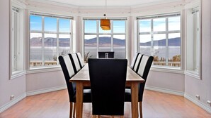 Een eetkamer met een grote tafel en stoelen, grote ramen met uitzicht op bergen en een wateroppervlak.
