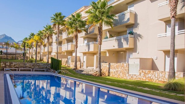 A swimming pool with clear blue water, surrounded by palm trees and a row of beige multi-story buildings with balconies.
