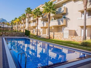 Una piscina con acqua cristallina azzurra, circondata da palme e da una fila di edifici beige a più piani con balconi.