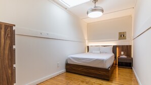 A bedroom with a wooden floor, a bed with white bedding, a skylight, and a bedside table with a lamp. - Suite 209: Beautiful 2-Bedroom Beachfront Suite (Wainfleet)