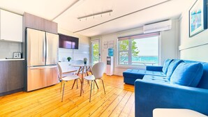 A modern kitchen with a built-in refrigerator, a dining area with a table and chairs, and a blue sofa in a living area with large windows. - Suite 209: Beautiful 2-Bedroom Beachfront Suite (Wainfleet)