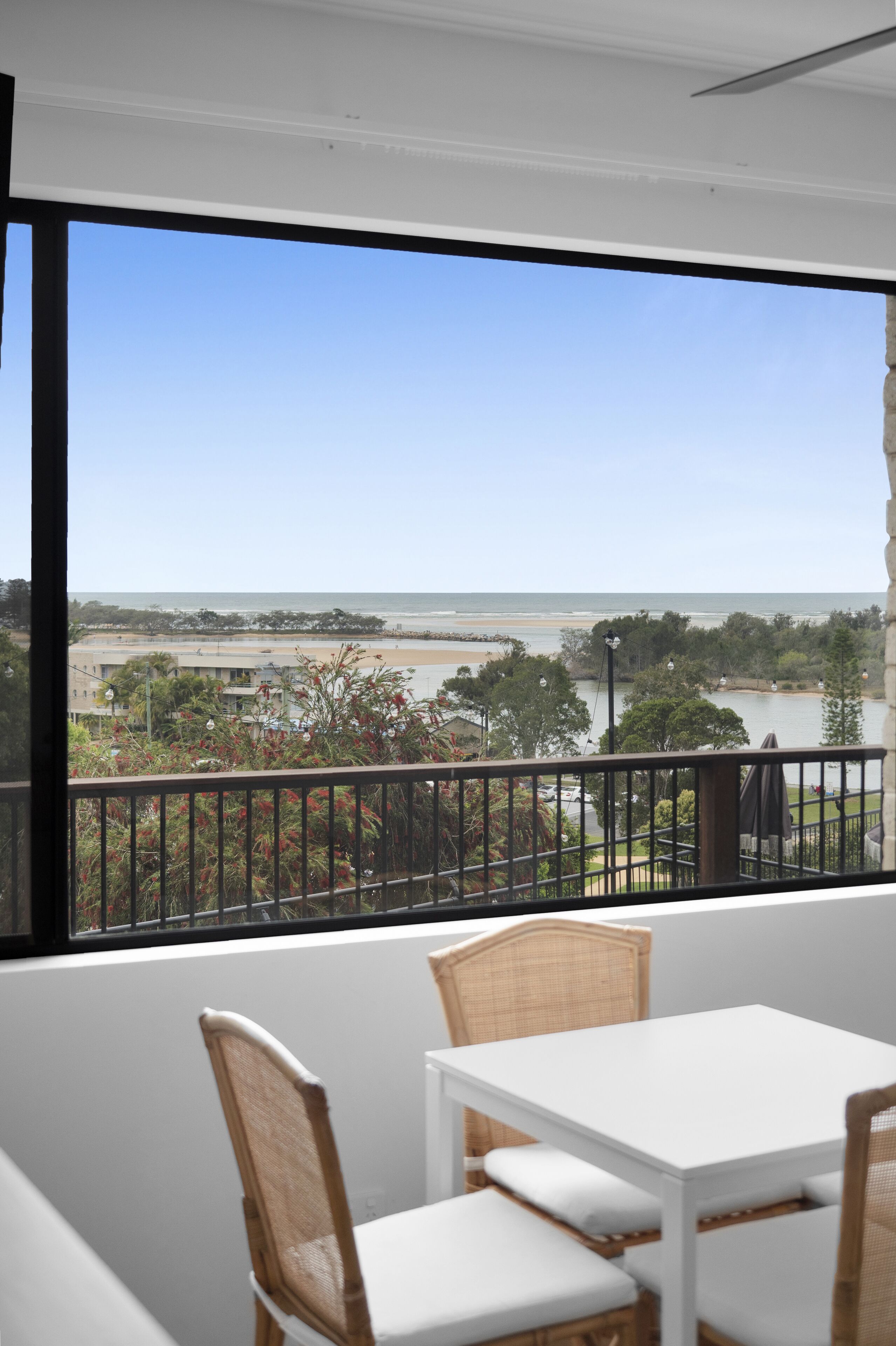 A balcony with a white table and wicker chairs, overlooking a beach and a body of water.