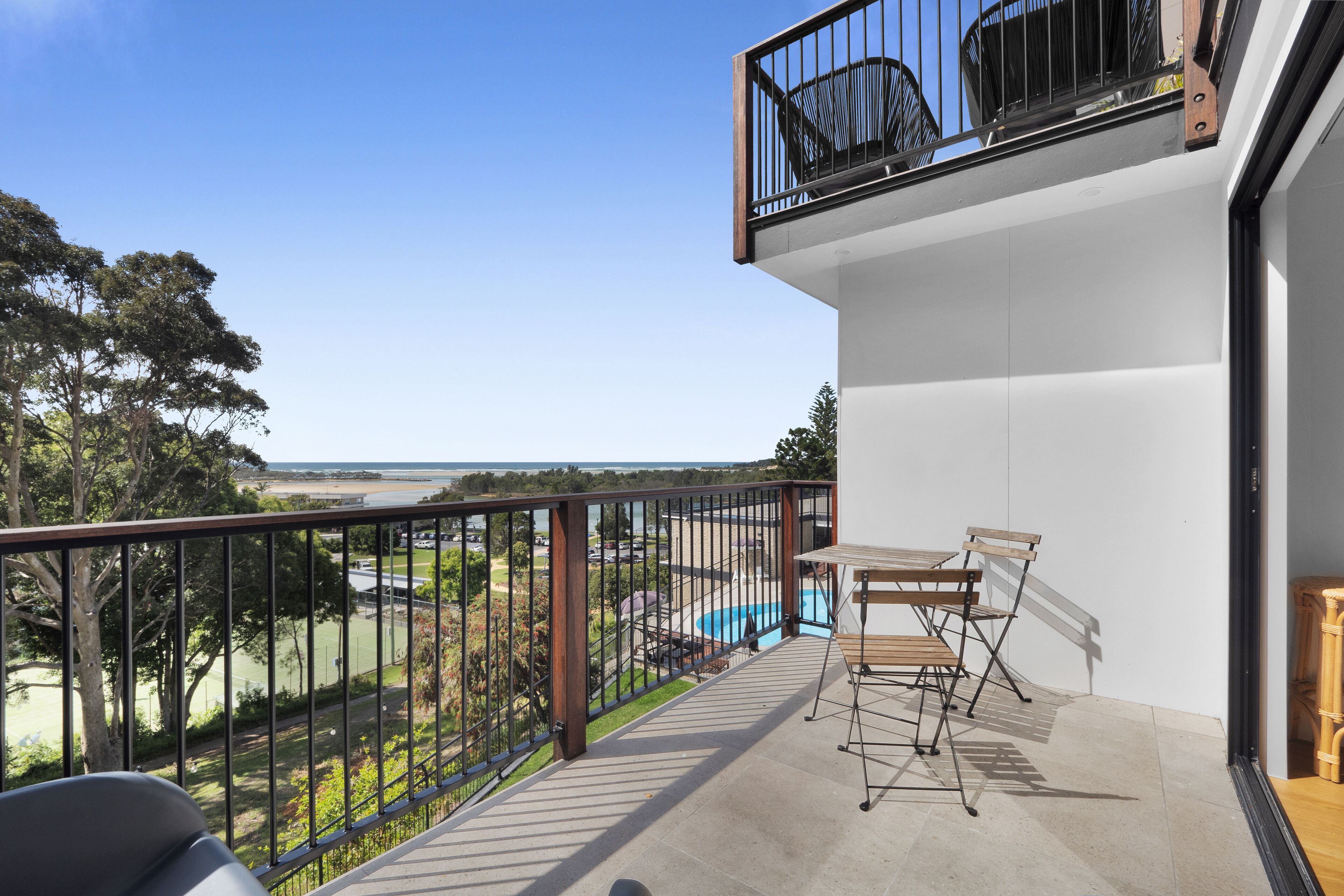A balcony with a view of a swimming pool and a tennis court, a table with chairs, and a wooden bench.