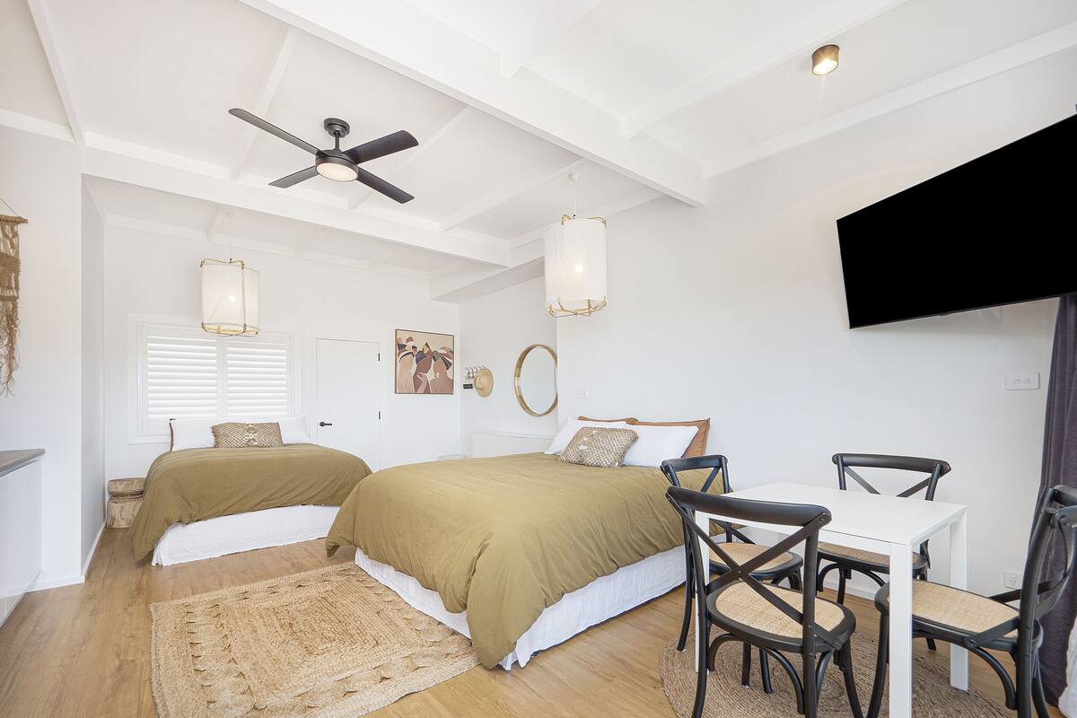 A bedroom with two beds, a ceiling fan, a TV, and a dining area with chairs and a table.
