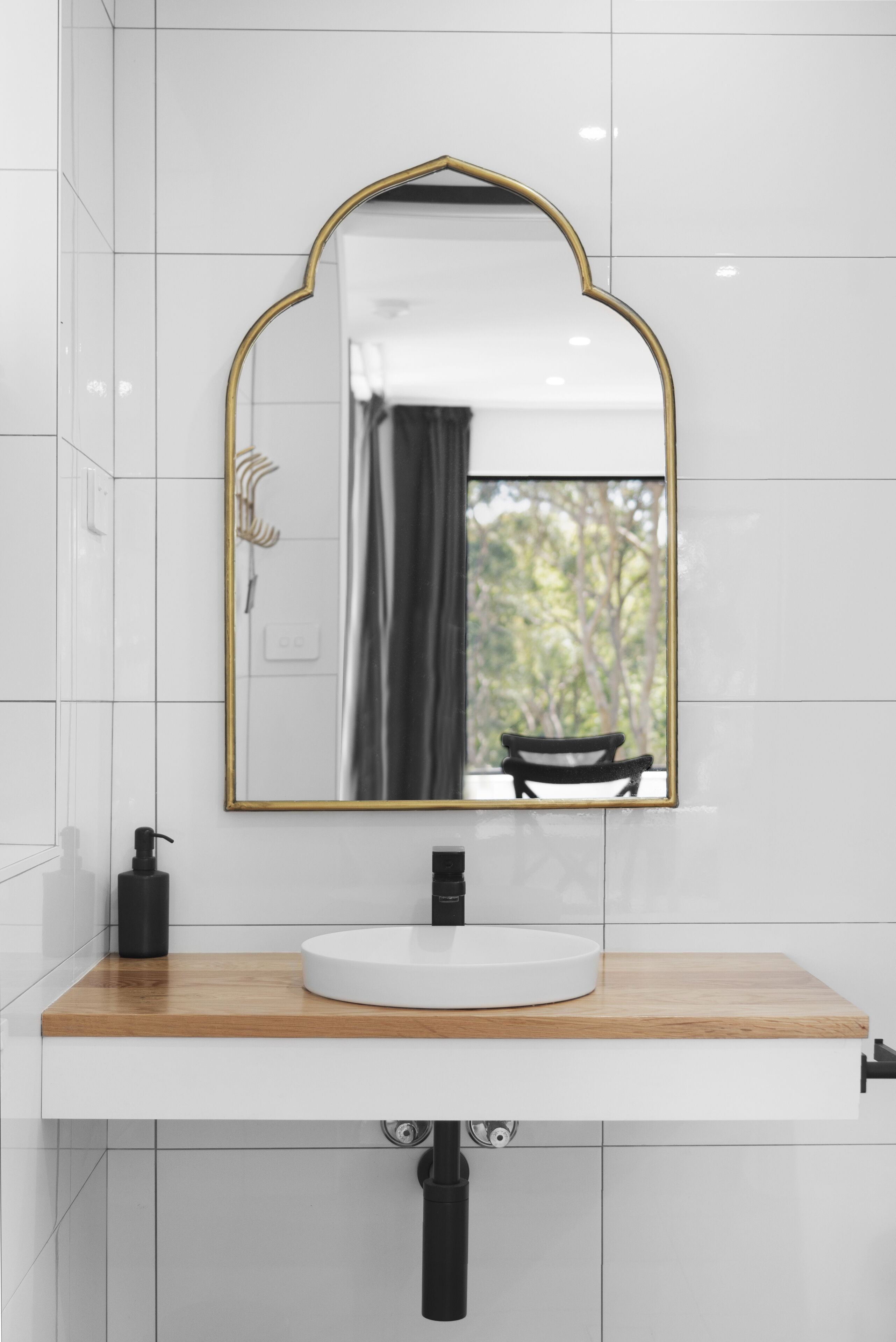 A bathroom with a white tiled wall, a wooden vanity, a white sink, a gold-framed mirror, and a black faucet.