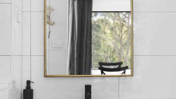 A bathroom with a white tiled wall, a wooden vanity, a white sink, a gold-framed mirror, and a black faucet.